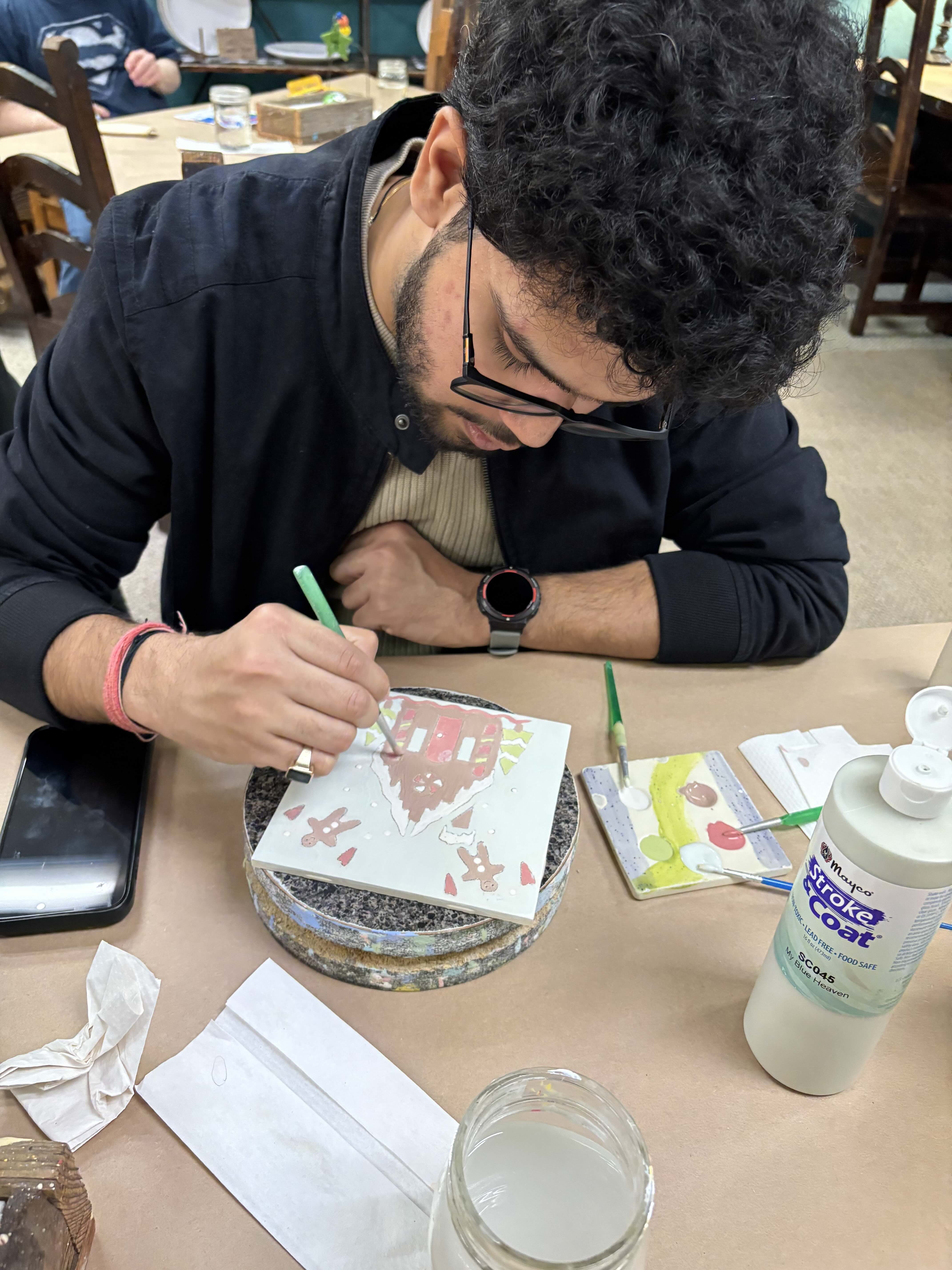 A young man concentrates on painting a colorful ceramic tile at a crafting table.